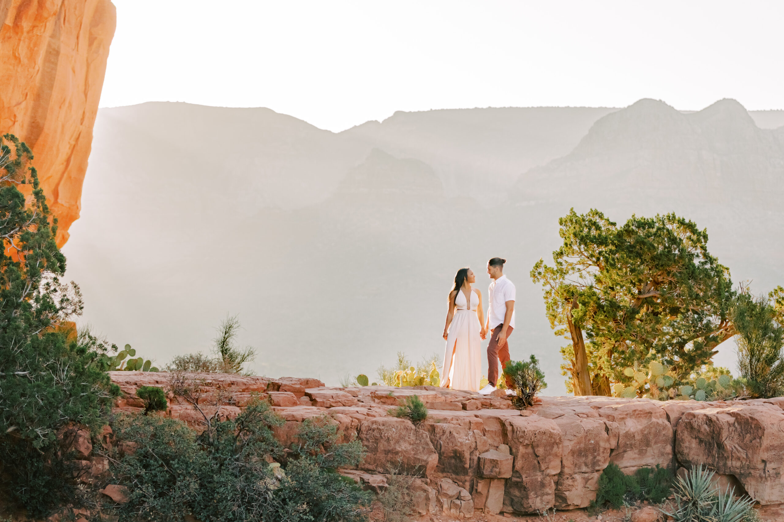 A newly engaged couple stands atop Sedona's beautiful red rocks. Behind them is a sunrise with light piercing through the cascading mountains, making the entire image illuminated from behind. The light embodies the happiness the couple is experiencing from being engaged and anticipating their wedding day.