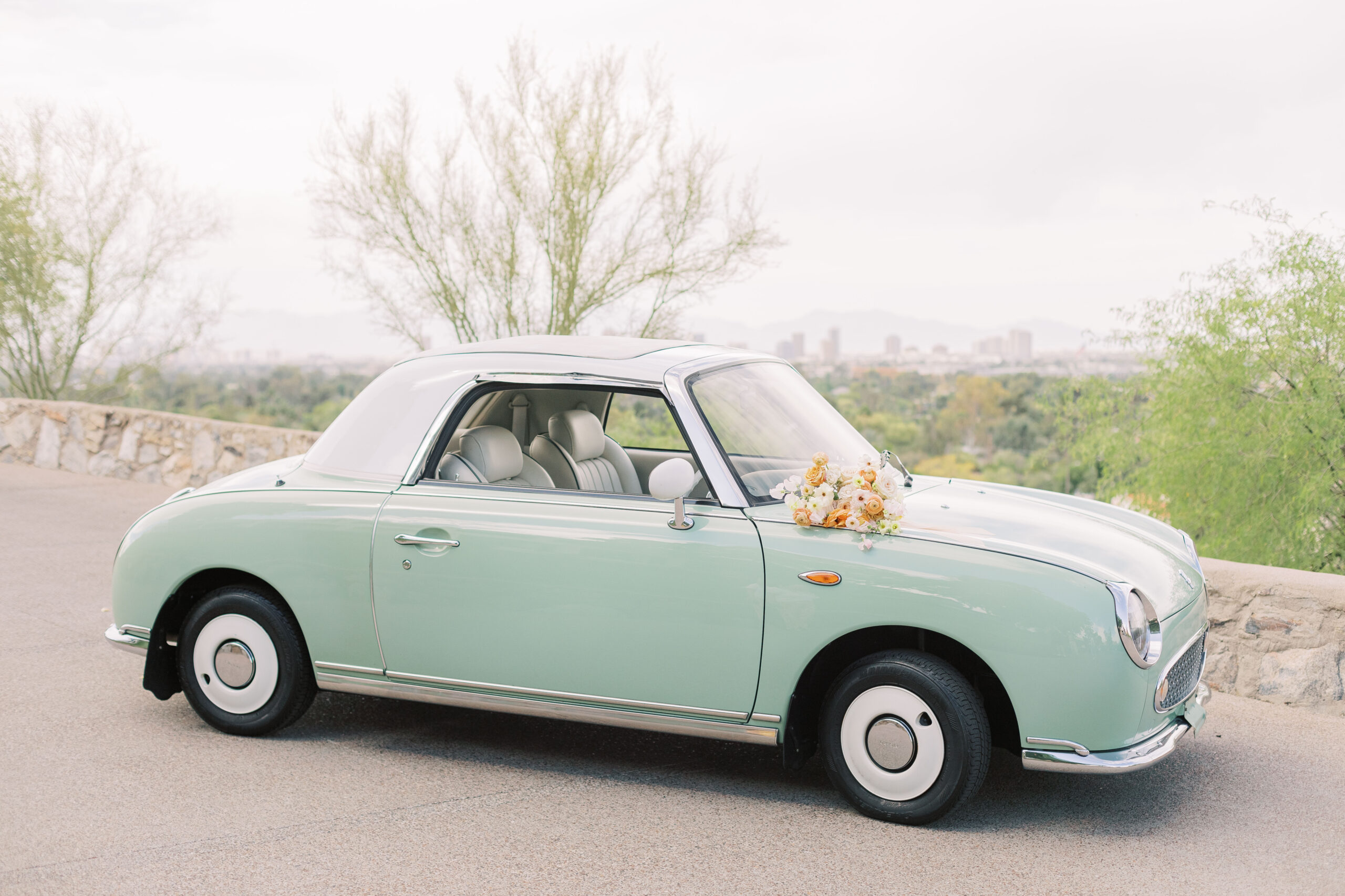 A cute vintage car sits atop a hill at one of Phoenix's famous wedding venues, Wrigley Mansion. The car is a beautiful mint color and it shows the bride's bouquet on the hood, which is filled with flowers that are orange, peach, and white. It is the car the bride and groom will be using when they exit the reception after celebrating their wedding day at Wrigley Mansion. Behind the car, is one of Wrigley Mansion's iconic views, which overlooks the entire city of Phoenix as well as Camelback Mountain. 