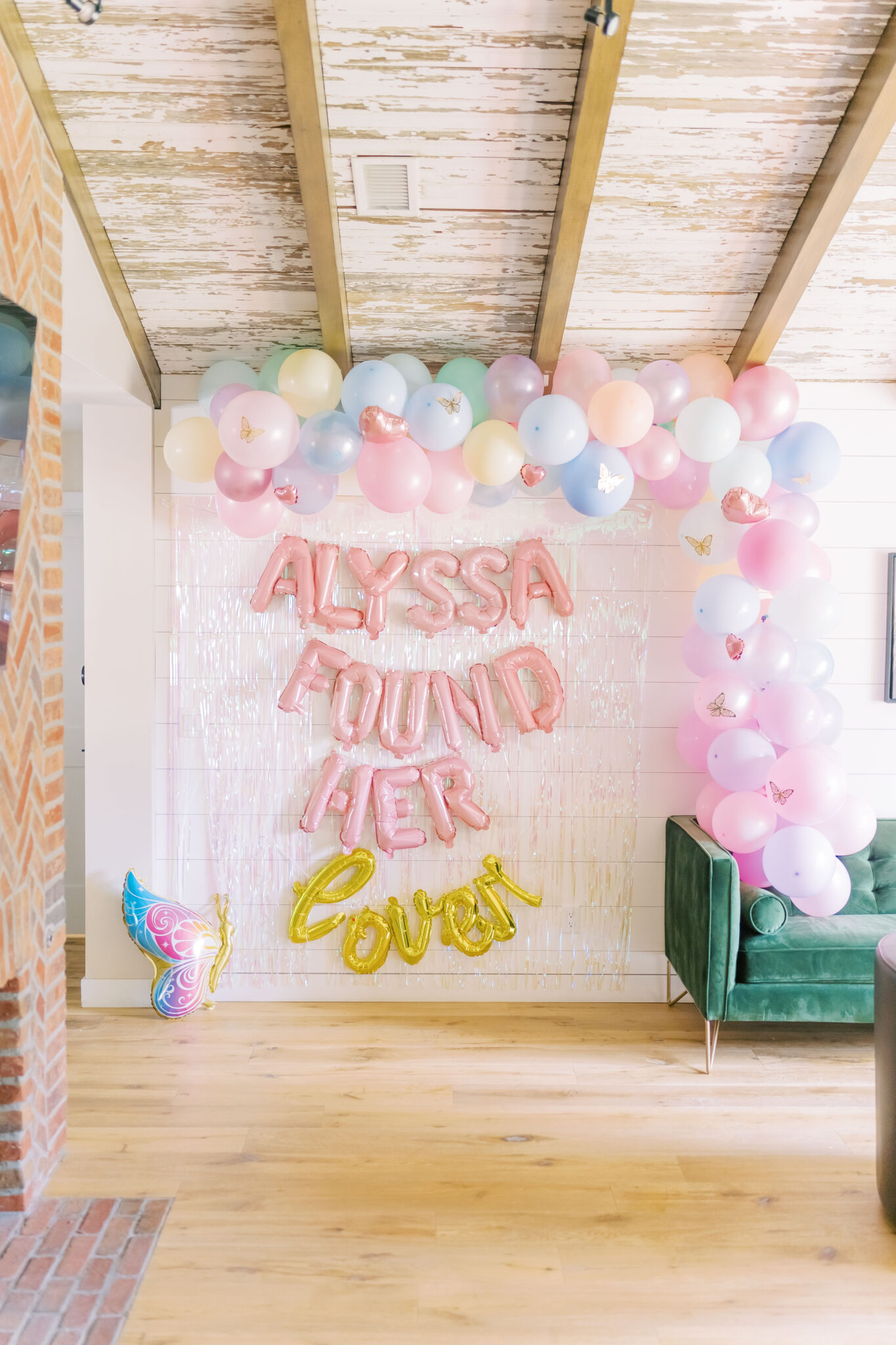 A wall is decorated with Taylor Swift themed decor like a balloon arch with pastel colored balloons and butterflies. In the middle, letter balloons spell out Alyssa Found Her Lover. 