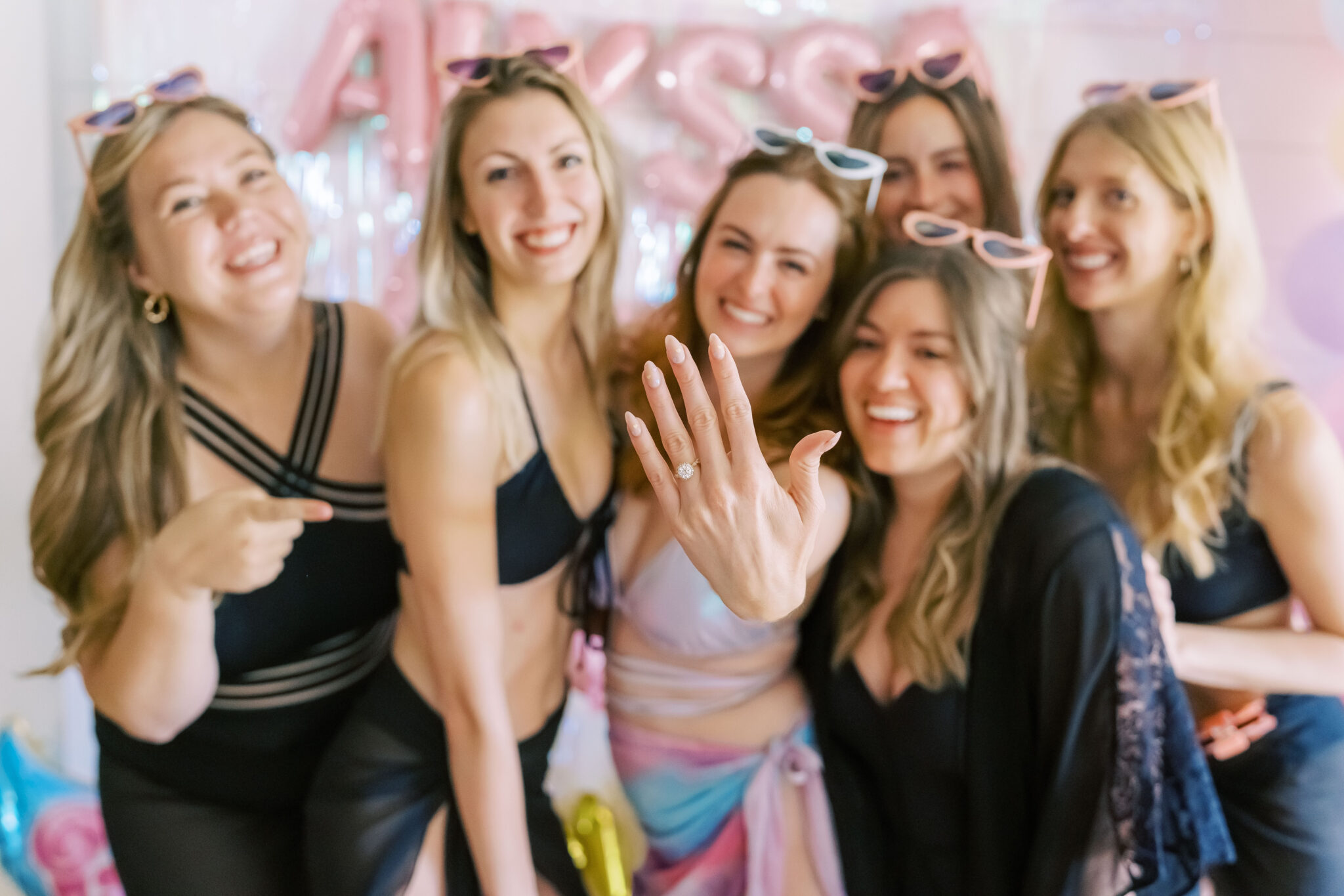 A group of five girls gather around the bride, showing off her engagement ring. They're standing in front of a decorated wall with balloons that spell out "Alyssa Found Her Lover." They are celebrating Alyssa's engagement with a Taylor Swift themed bachelorette party in Scottsdale, Arizona.